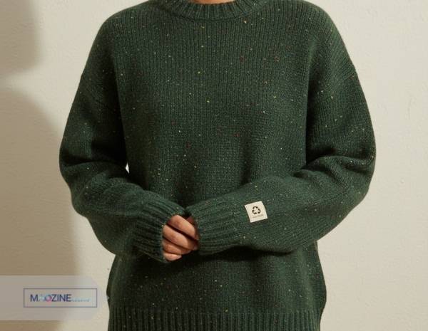A person wearing a stylish, dark green sustainable fashion sweater made from recycled wool, with a small recycled symbol logo on the cuff.