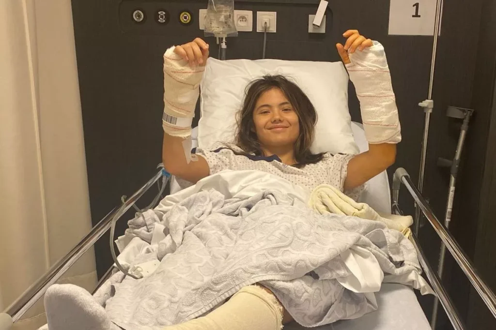 Emma Raducanu smiling in a hospital bed with bandaged wrists and ankle after undergoing triple surgery in 2023.