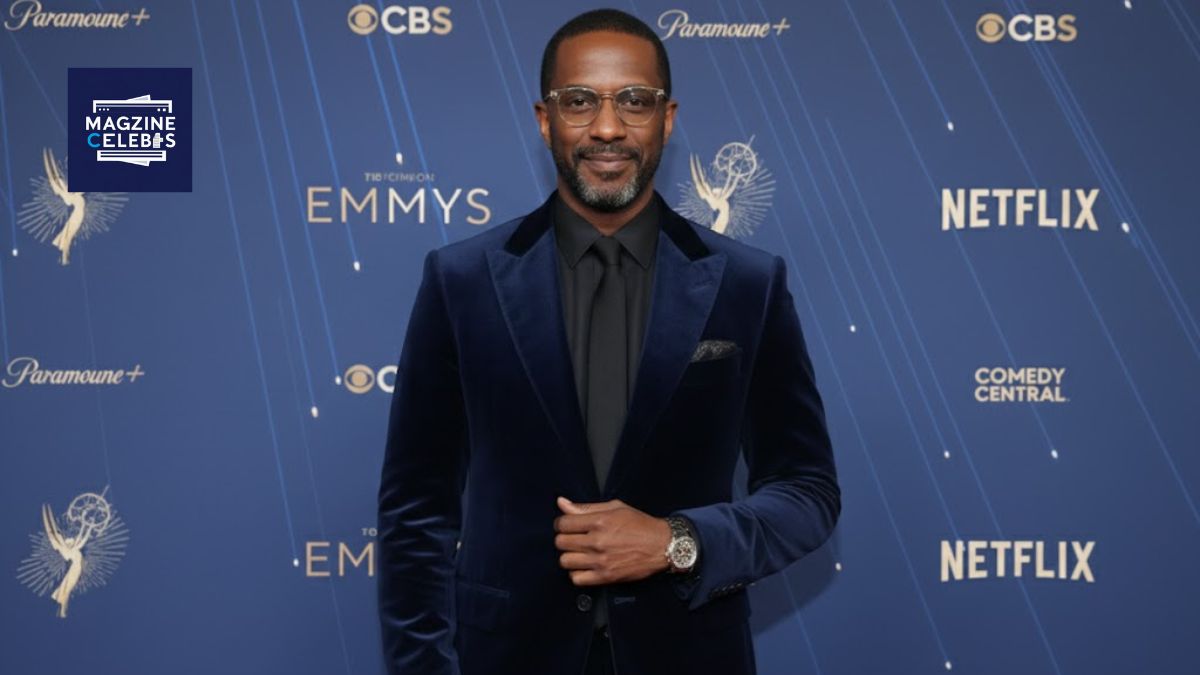 A portrait of comedian Dave Chappelle wearing a suit and glasses, posing in front of a media wall at a 2025 event.