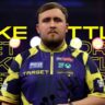 Luke Littler Net Worth 2025 – The Nuke holding darts on stage in yellow and blue Target Darts jersey during World Darts Championship