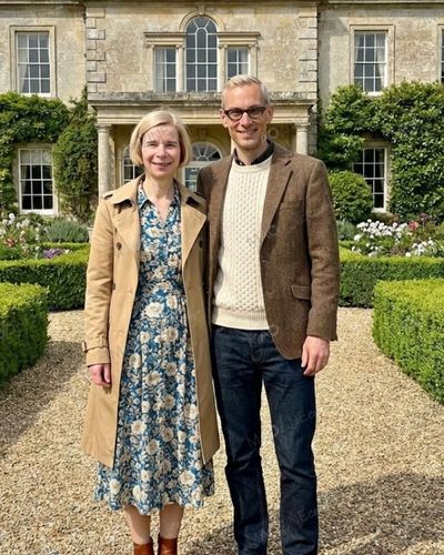 Architect Mark Hines standing next to his wife Lucy Worsley in front of a grand historic manor. Mark is wearing a tweed jacket and glasses, reflecting his work in heritage conservation.