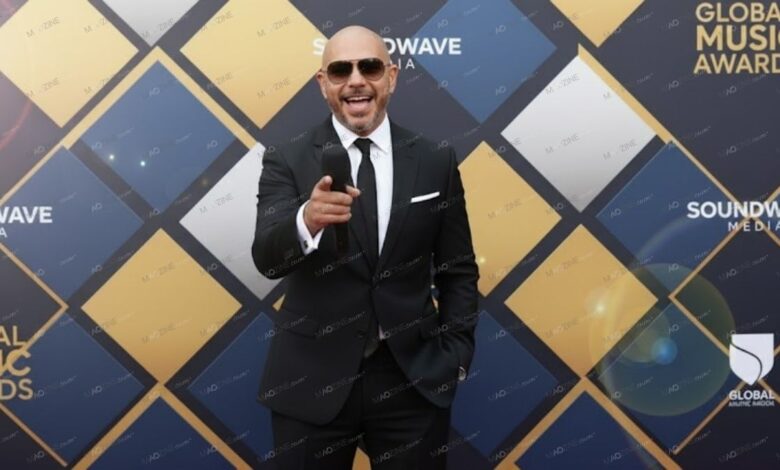 Pitbull wearing a suit and sunglasses, pointing at the camera while holding a microphone on the red carpet at the Global Music Awards.